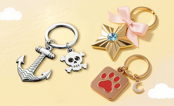 Shiny metal anchor and skull keychains, gold star charm with blue gemstone, pink ribbon, paw print charm, and crescent moon charm with cozy background.