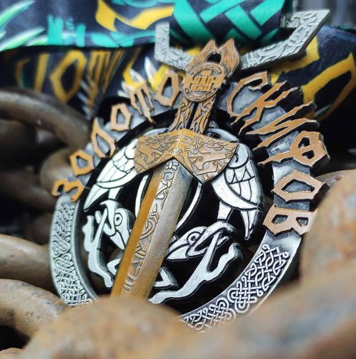 Medal featuring Norse mythology design, bronze and silver accents, awarded on a colorful ribbon, lying on rusty chain links.